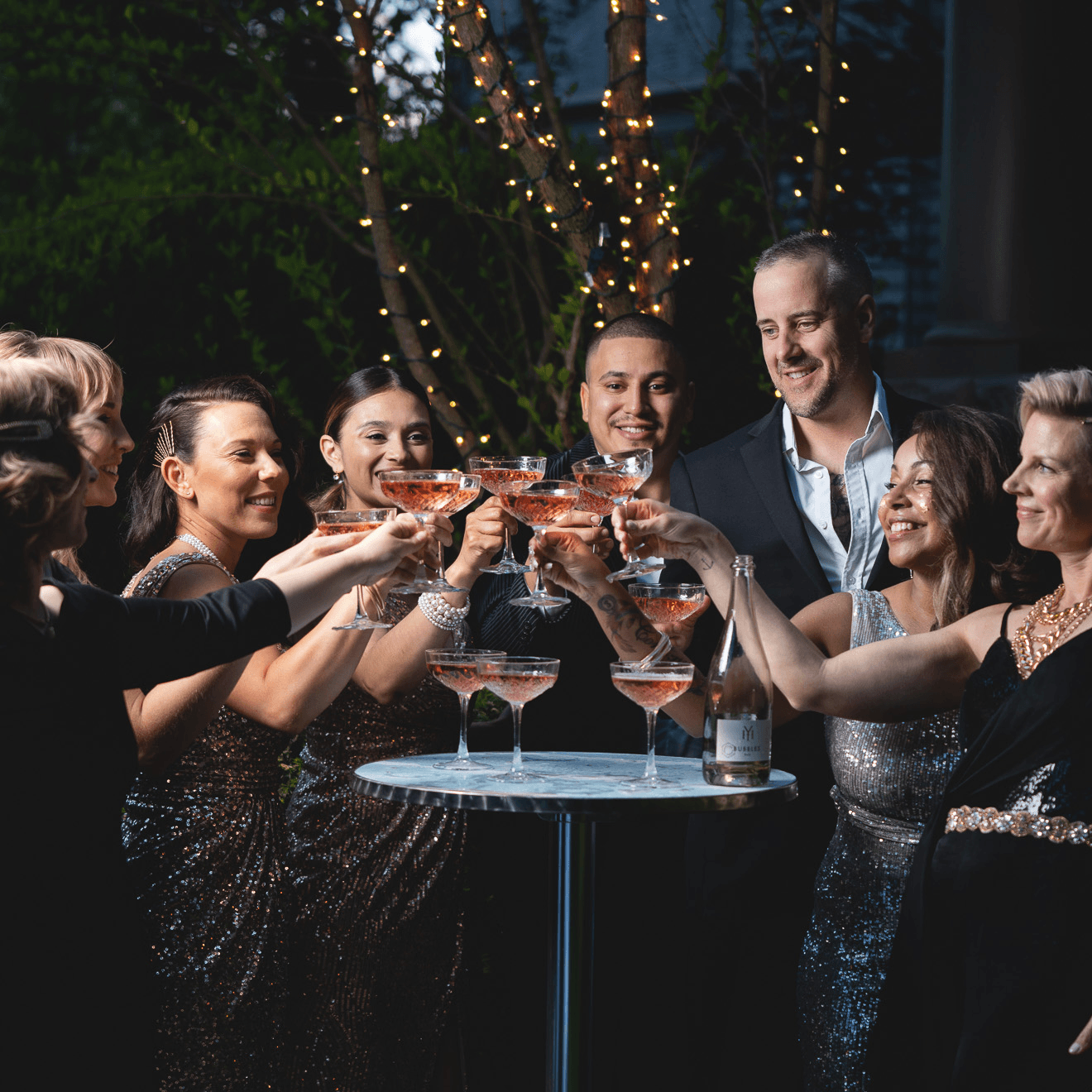 Group of people all holding champagne glasses filled with pink champagne in the center of the group, to toast