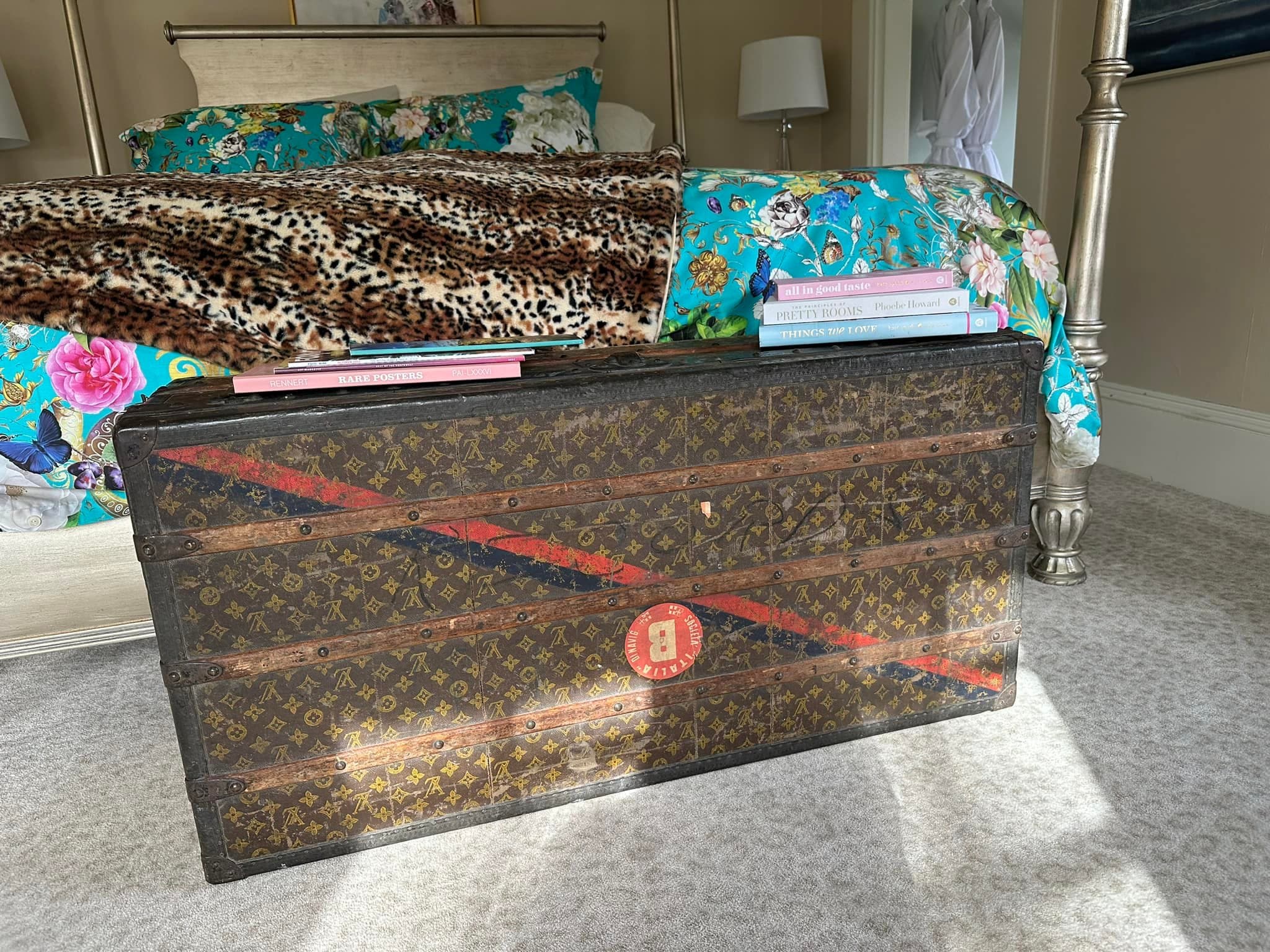 A vintage Louis Vuitton trunk sits at the foot of a bed adorned with colorful bedding and books on top.