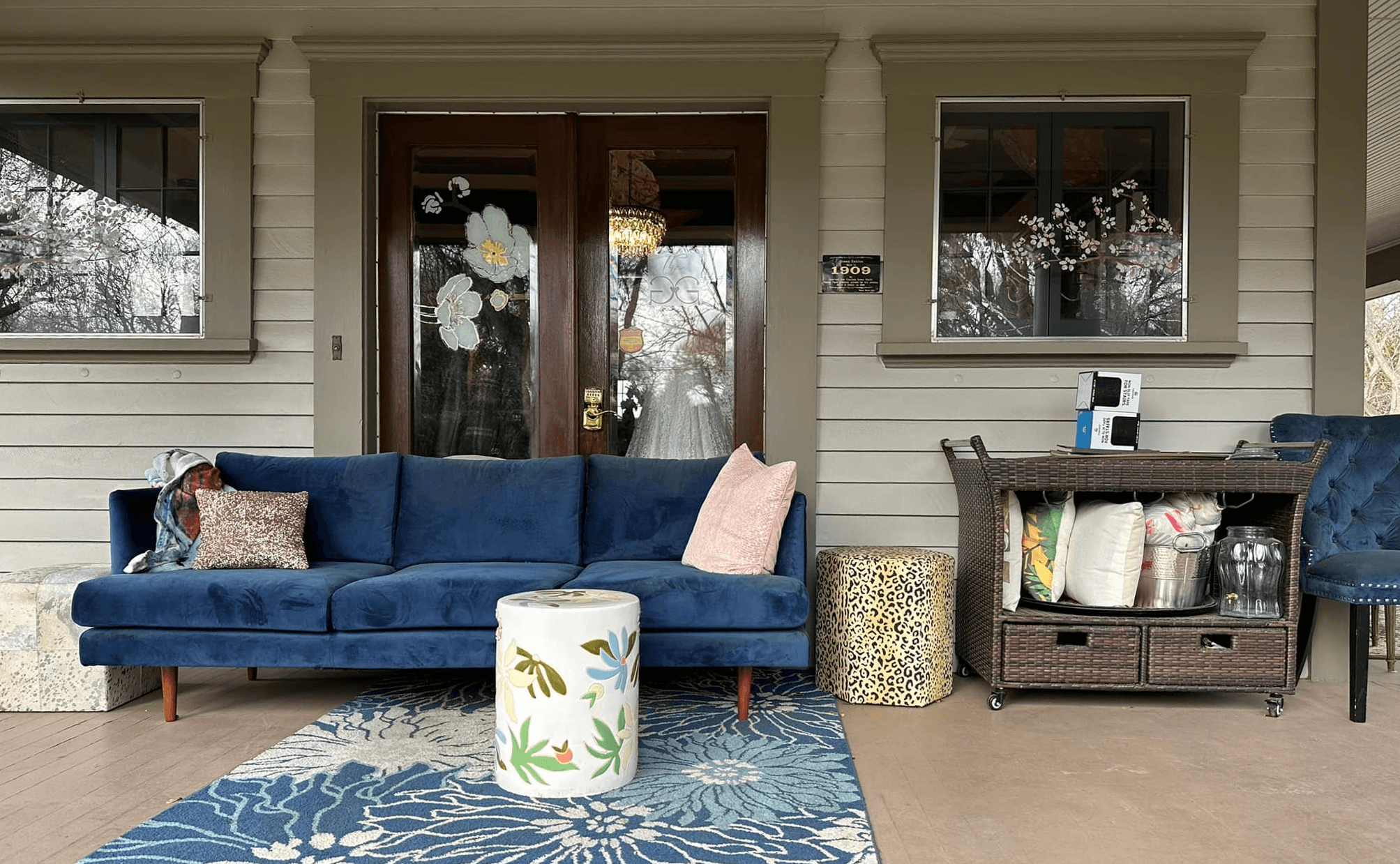 A cozy porch featuring a blue sofa, decorative stools, and a cart with pillows and blankets.