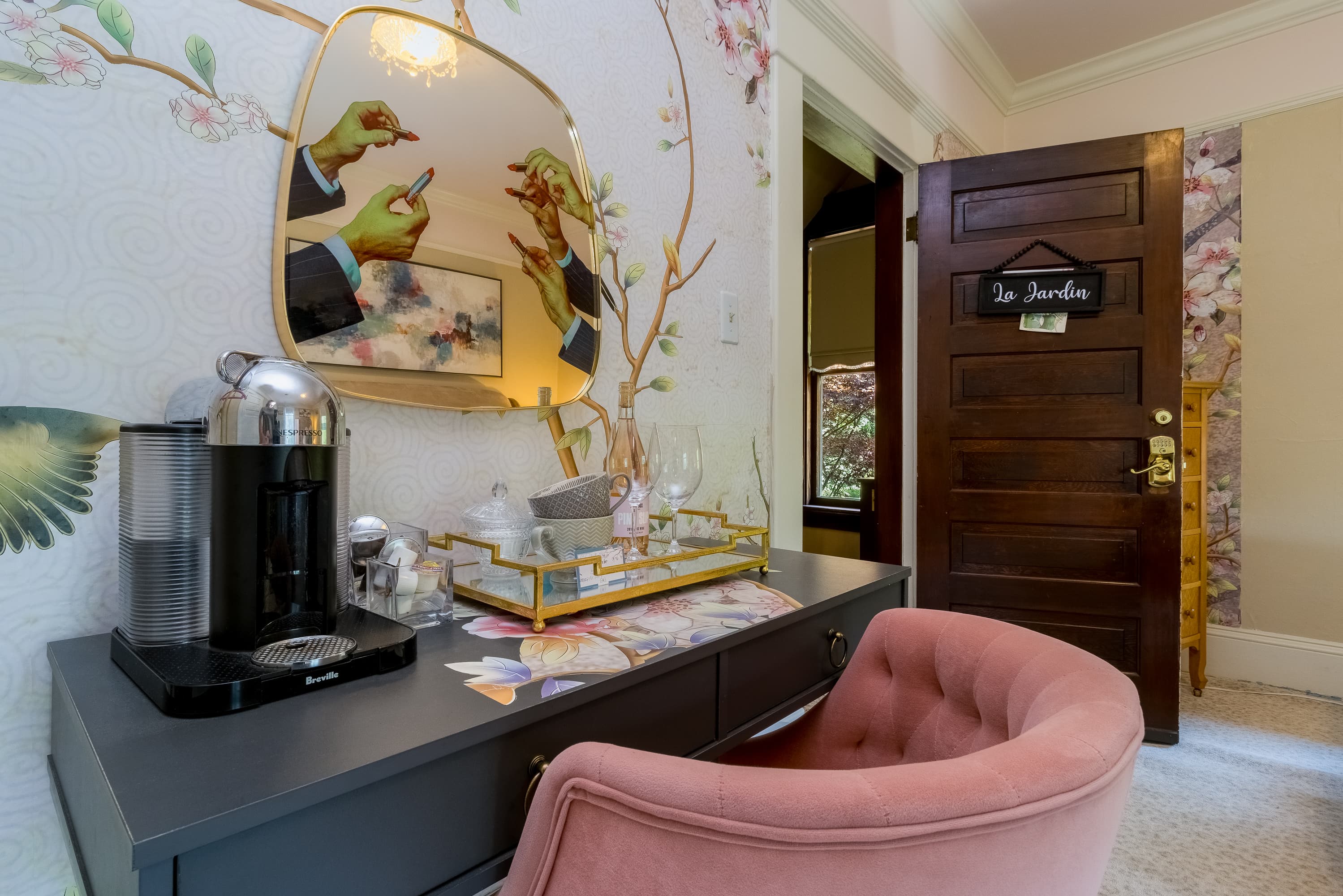 A chic vanity area with a pink chair, coffee maker, and floral decor, highlighted by a mirror reflecting artistic hands.