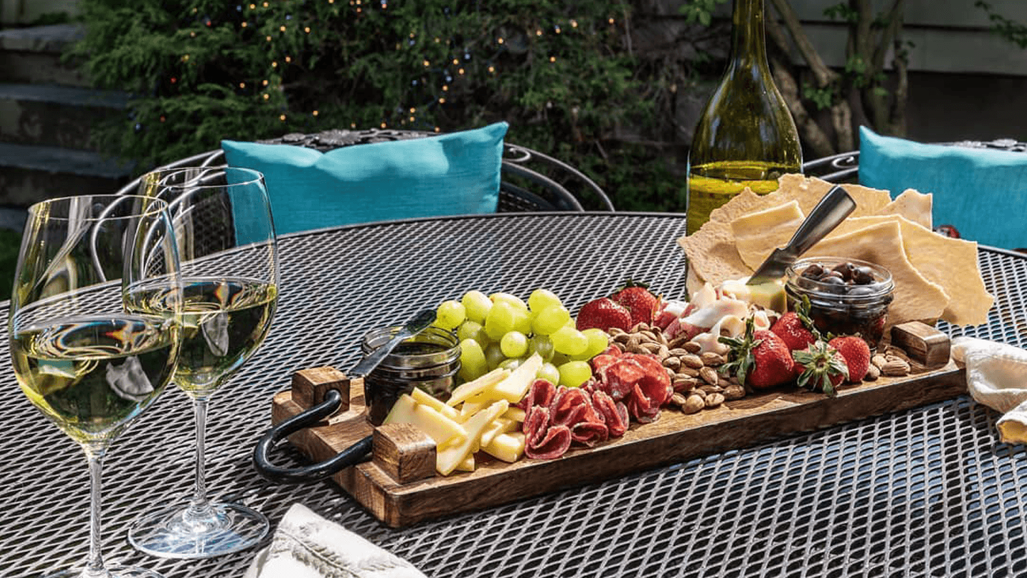 A wooden platter with various cheeses, meats, grapes, strawberries, nuts, and a jar of olives, accompanied by two glasses of white wine, set on a table.