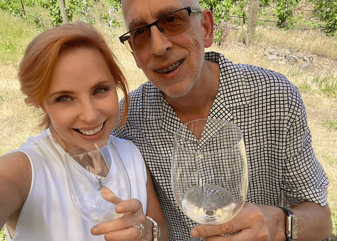 Karrie and Brian holding glasses of white wine, taking a selfie at a vineyard