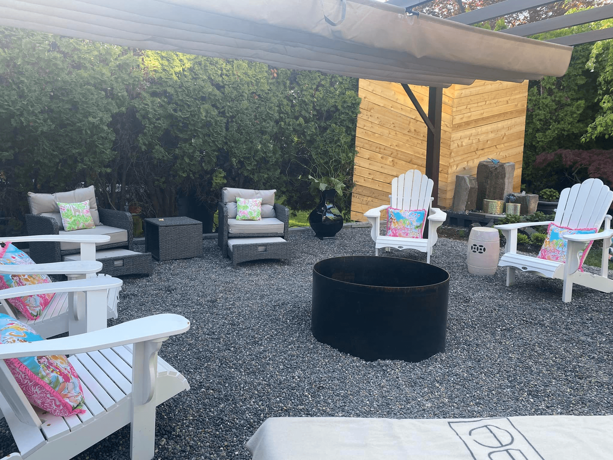 A cozy outdoor seating area with white and wicker chairs, colorful cushions, and a fire pit surrounded by gravel and greenery.