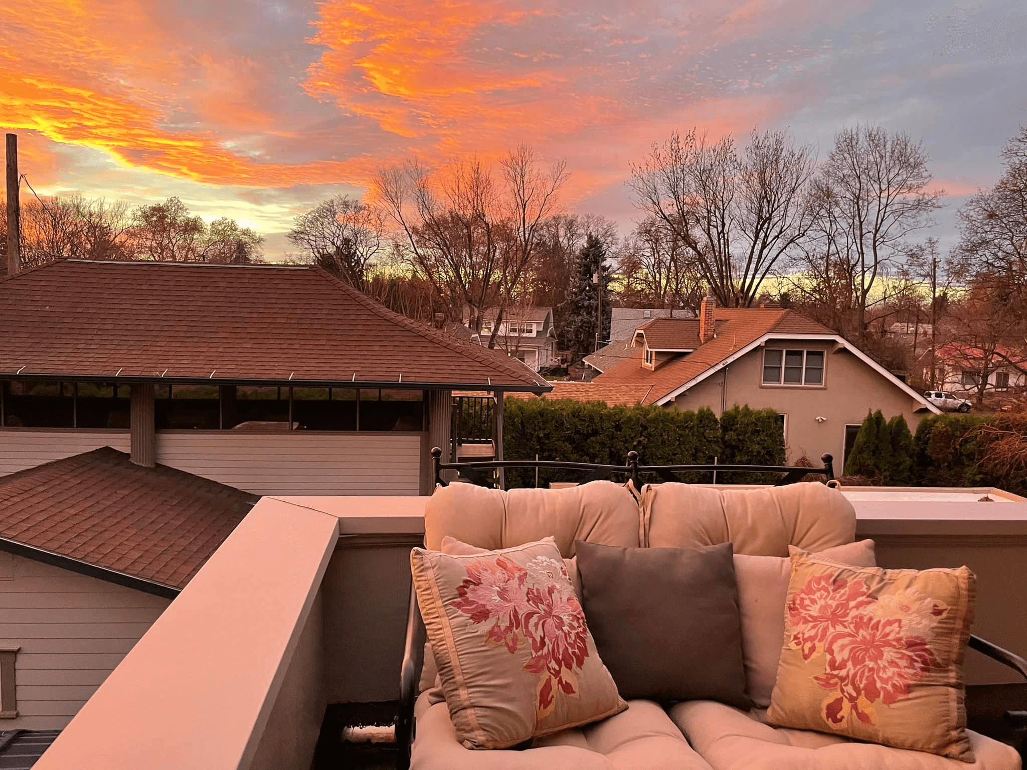 A cozy balcony seating area with floral pillows overlooks rooftops against a vibrant sunset sky.