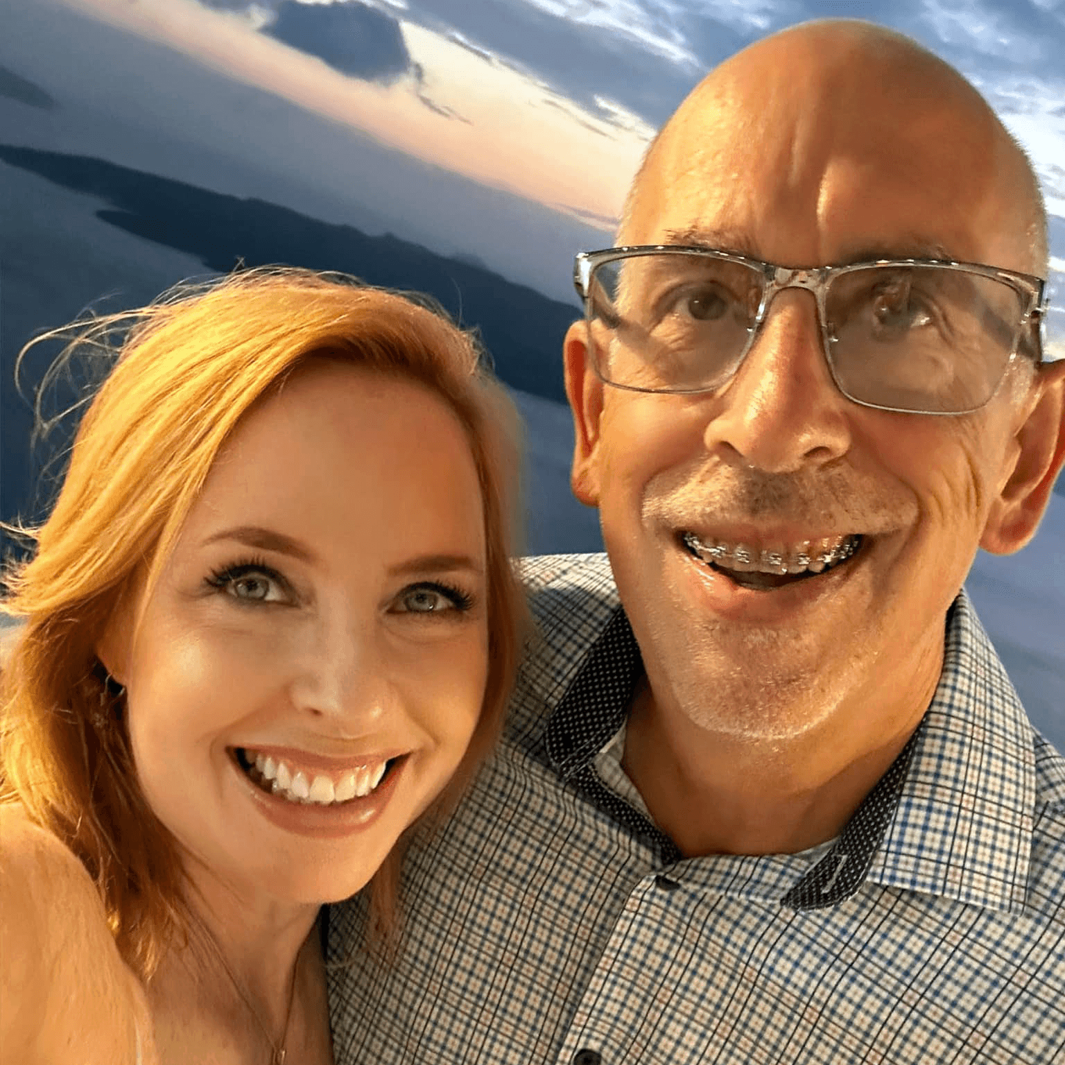 A man and a woman smile together in a selfie against a scenic sunset background.