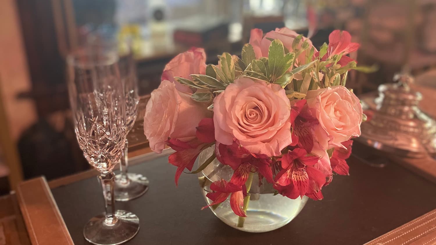Bowl of pink, large rose blooms next to a pair of crystal champagne flutes