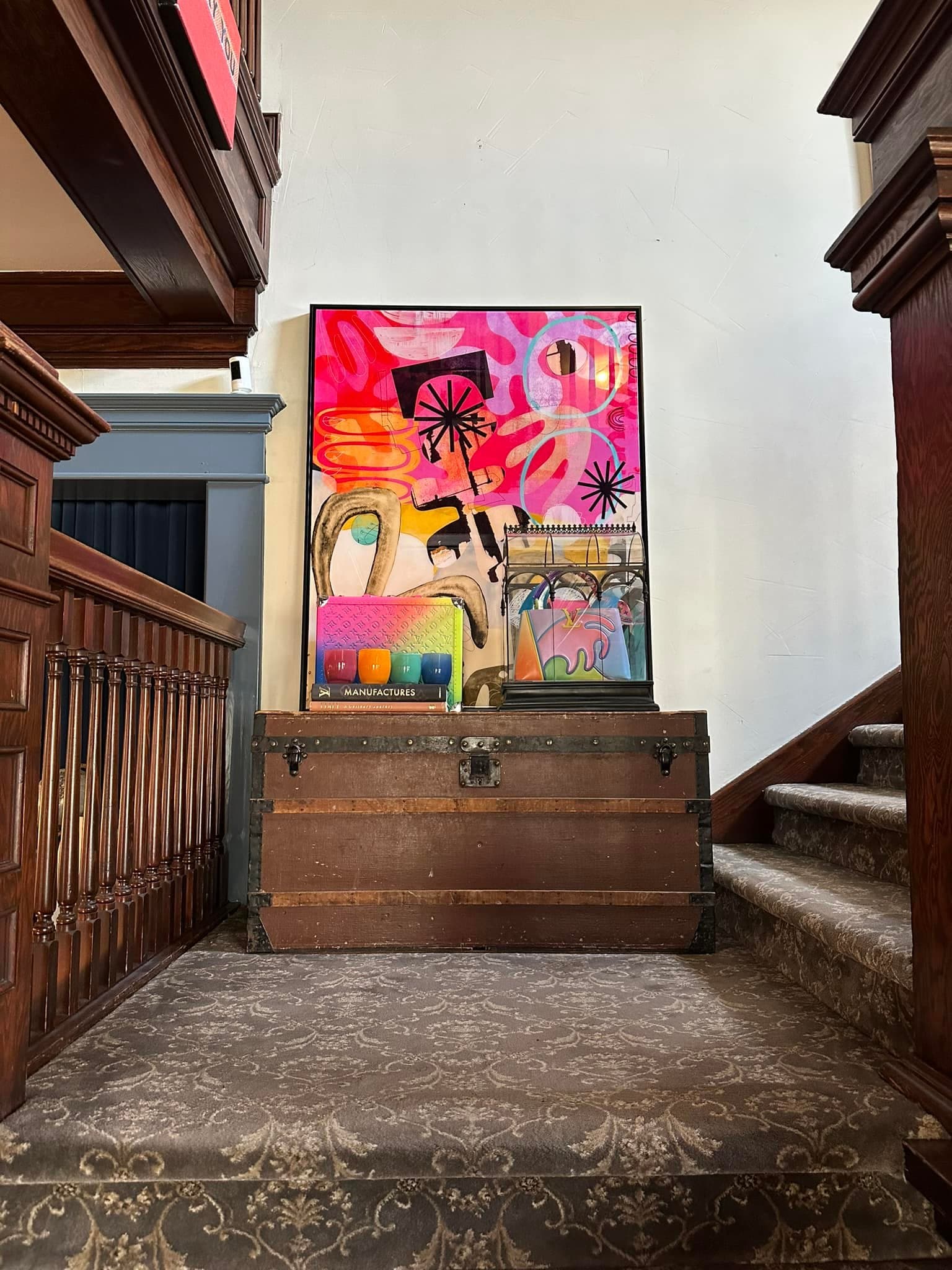 A colorful abstract painting is displayed above a wooden trunk on a staircase.