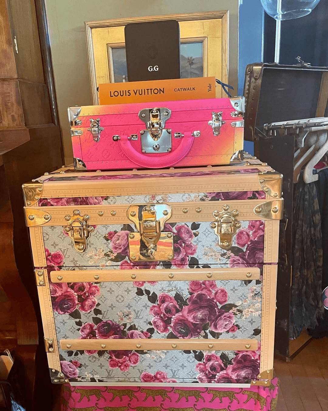 A stack of decorative luggage, including a floral-patterned trunk and a pink case, rests in a room with a framed picture in the background.
