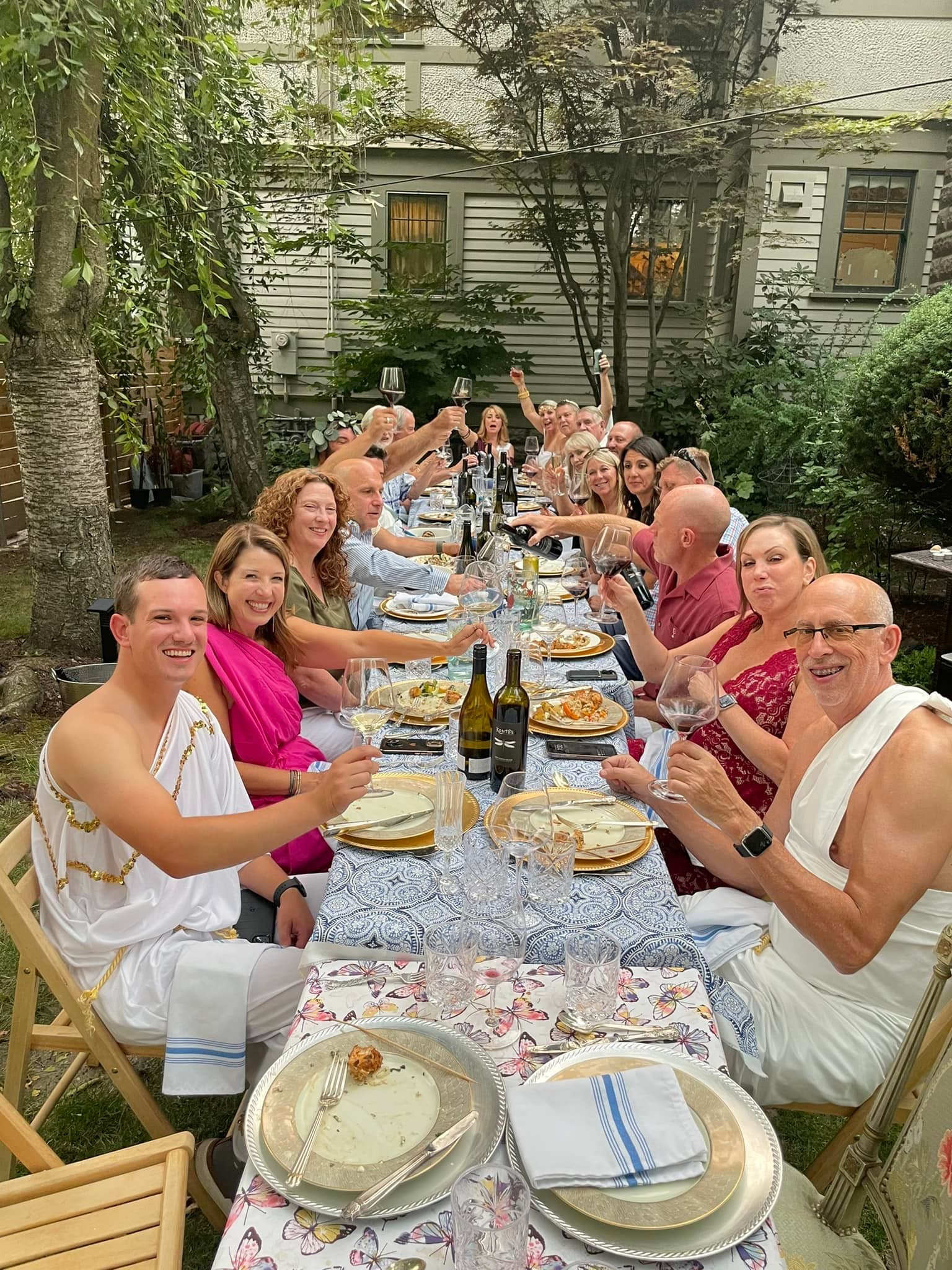 A festive outdoor gathering with guests in togas enjoying a meal and raising glasses in celebration.