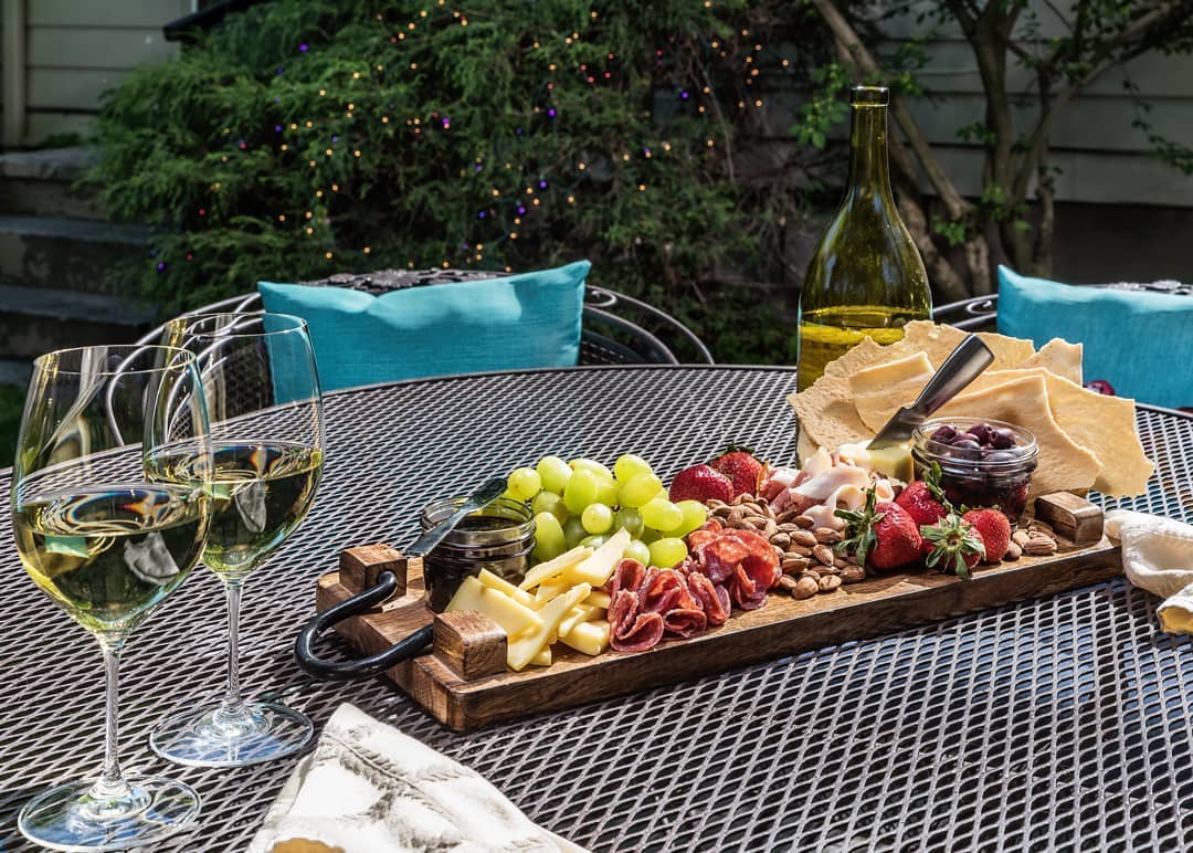 A wooden platter filled with assorted cheeses, meats, fruits, and nuts is set on a table alongside two glasses of white wine.