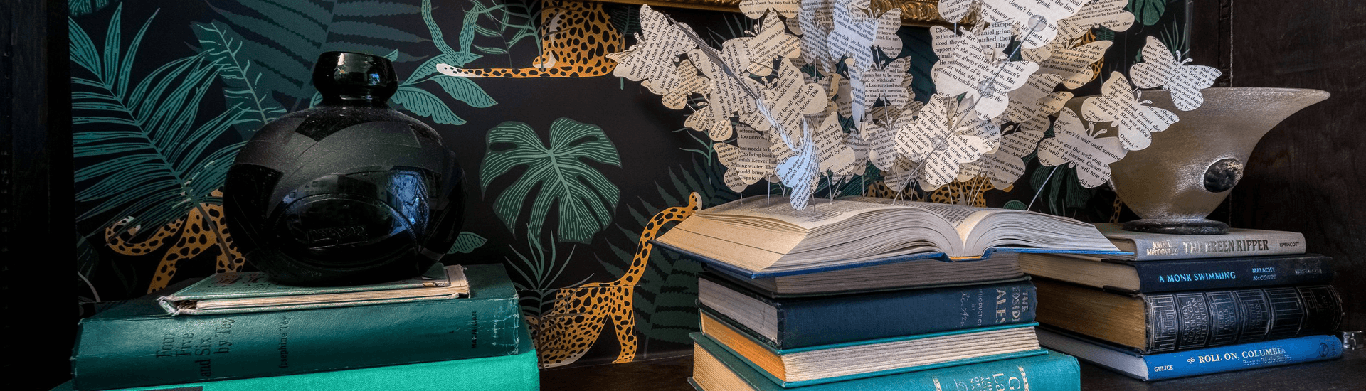 A stack of vintage books beside a decorative glass vase and a paper butterfly sculpture against a patterned background.
