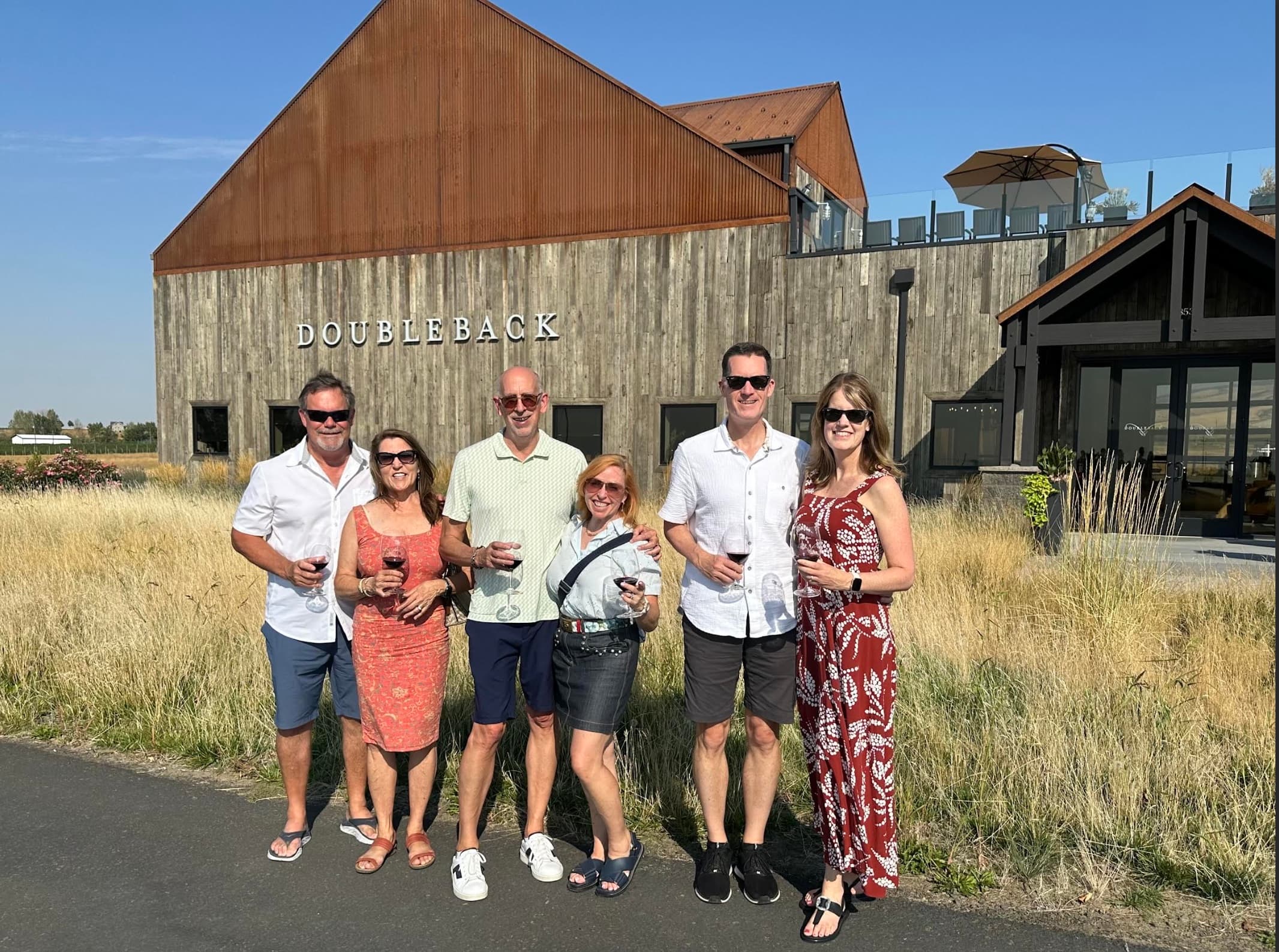 A group of six smiling guests holding glasses of red wine outside Doubleback Winery in Walla Walla, enjoying a sunny day during a curated retreat experience. A group of six smiling guests holding glasses of red wine outside Doubleback Winery in Walla Walla, enjoying a sunny day during a curated retreat experience.